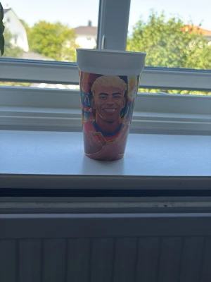 mcdonalds lamine yamal mugg - Säljer en färgglad plastmugg med ett tryckt porträtt av en leende person med blont hår. Muggen har vit kant upptill och motivet är i starka färger. Perfekt för drycker hemma eller på utflykt.