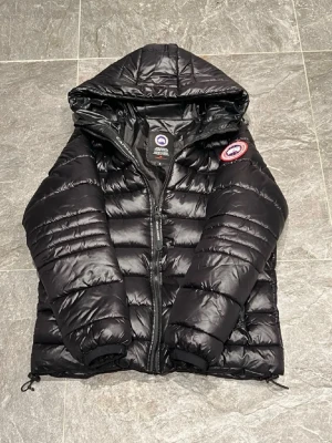 Svart dunjacka från Canada Goose - Säljer en svart dunjacka från Canada Goose med huva och dragkedja. Jackan har det klassiska runda Canada Goose-märket på ärmen och är tillverkad i glansigt material med quiltad design. Perfekt för kalla vinterdagar.