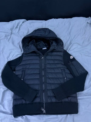 Moncler cardigan  - Snygg svart pufferjacka från Moncler med huva och stickade ärmar. Jackan har dragkedja framtill, ribbade muddar och fickor med knappar. Moncler-logga på ena ärmen och coola detaljer som zipficka på armen. Perfekt för kalla vinterdagar.