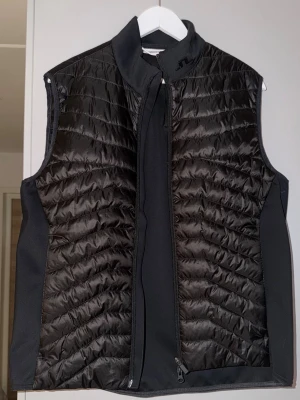 Svart dunväst från J.Lindeberg - Snygg svart dunväst från J.Lindeberg med hög krage och diskret logga på kragen. Västen har quiltad design framtill och släta sidopaneler i stretchigt material. Perfekt lager på lager och stängs med dragkedja framtill.