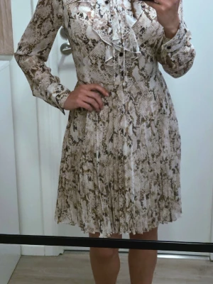 Kort klänning med ormmönster och volang från Karen Millen - Säljer en kort klänning med beige och brunt ormmönster. Klänningen har långa ärmar, knappar framtill och volangdetalj vid halsen. Materialet känns lätt och luftigt, perfekt för dig som gillar statementplagg med coola detaljer. Sparsamt använd och inga synliga fläckar. St 10 (brittisk) eller 38 (EU) men är nog mer st 36 i passformen 