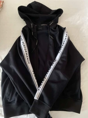 Svart hoodie Peak Performance - Svart hoodie från Peak Performance med vita band längs ärmarna där märkesnamnet står tryckt. Klassisk huva med dragsko och ribbade muddar vid ärmslut och nederkant. Perfekt för en avslappnad och sportig stil.