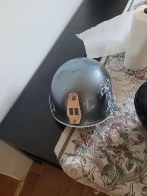 Grå hjälm med vita blommor - Säljer en grå hjälm med vita blommönster på sidorna. Hjälmen har en unik design och detaljer i beige framtill. Passar perfekt för dig som vill sticka ut med din utrustning.