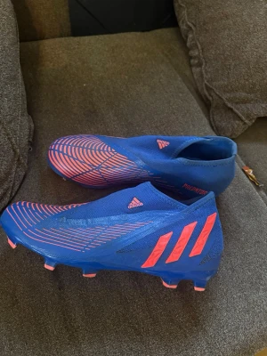 Blåa Adidas Predator fotbollsskor - Säljer ett par blåa Adidas Predator fotbollsskor med rosa detaljer och mönster. Skorna har en strumpliknande passform utan snörning och är designade för spel på konstgräs eller gräs. Perfekta för dig som vill ha kontroll och stil på planen.