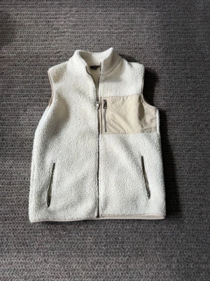 Beige teddyväst med dragkedja - Snygg vit teddyväst i mjukt, fluffigt material med hög krage och hel dragkedja framtill. Västen har två sidofickor med dragkedja och en beige bröstficka i nylon med dragkedja för extra stil. Perfekt för lager på lager och trendig streetwear-look.