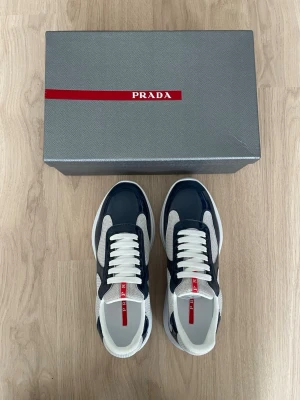 Prada cups i marinblå och vit - Snygga Prada sneakers i marinblått och vitt med röda detaljer. Skorna har en chunky vit sula, meshpaneler och lackade partier i läder. Klassisk låg modell med vita skosnören och tydlig Prada-logga på plösen.