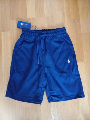 Marinblå shorts från Polo Ralph Lauren - Snygga marinblå shorts från Polo Ralph Lauren med klassisk vit logga broderad på benet. Shortsen har resårmidja med snörning och två sidofickor. Perfekta för sommardagar och avslappnade tillfällen.