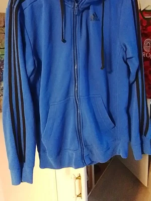 Adidas hoodie, dragkedja - Blå hoodie från Adidas med svarta ränder längs ärmarna och dragkedja framtill. Klassisk huva med svarta snören och två stora fickor på magen. Adidas-logga på bröstet. Perfekt för en sportig och avslappnad stil.