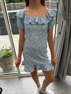 Blå blommig kortklänning med volang - Modellen är 157. Supersöt ljusblå kortklänning med små vita blommor, volangdetaljer vid axlar och fåll samt korta puffärmar. Klänningen har en feminin och somrig vibe, perfekt för varma dagar. Materialet känns lätt och luftigt.