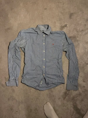 Blårutig långärmad skjorta - Säljer en klassisk blårutig skjorta med lång ärm och knappar framtill. Skjortan har en vit och blå rutig design och en liten broderad detalj på bröstet. Perfekt till jeans eller chinos för en clean look.