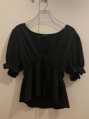 Svart blus med puffärm från Gina Tricot - Trendig svart blus från Gina Tricot med puffiga korta ärmar och volangdetaljer vid ärmslut. Blusen har en djup v-ringning och knytband framtill som ger en snygg siluett. Tillverkad i ett mjukt, stretchigt tyg som sitter bekvämt.