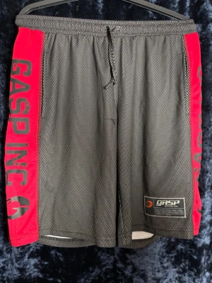 GASP INC shorts med röda sidor - Snygga träningsshorts från GASP INC med svart meshmaterial och röda paneler på sidorna. Stora logotexter längs benen och justerbar midja med snörning. Perfekta för gymmet eller andra sportaktiviteter. Skickas snabbt inom 1-2 dagar. Djur- och rökfritt hem. 
