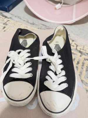 Svarta låga sneakers med vita detaljer - Klassiska svarta låga sneakers med vit tåhätta och vita skosnören. Skorna har en vit sula med svart rand och är tillverkade i canvasmaterial. Perfekta för en avslappnad och stilren look.
