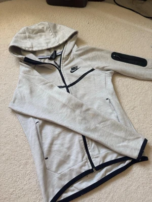 Ljusgrå Nike hoodie med svarta detaljer - Snygg ljusgrå hoodie från Nike med svarta detaljer och dragkedja. Se fläck på sista bild, inget man tänker på alls! Har ej testat att tvätta 