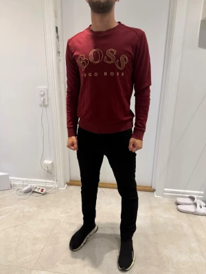 Vinröd BOSS sweatshirt - Snygg vinröd sweatshirt från Hugo Boss med stort BOSS-tryck i guld på bröstet. Tröjan har rund halsringning, långa ärmar och ribbade muddar. Perfekt för en stilren och avslappnad look.