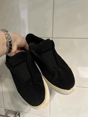 Svarta sneakers från Axel Arigato - Stilrena svarta sneakers från Axel Arigato i mocka med vit sula och diskret guldfärgad logga på sidan. Slip-on modell utan snörning för en clean look. Passar perfekt till streetwear och casual outfits.
