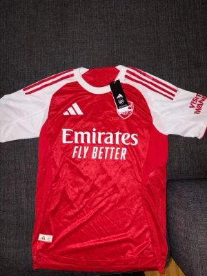 Arsenal matchtröja röd/vit Adidas - Snygg Arsenal matchtröja från Adidas i rött och vitt med klubbmärke och sponsortryck på bröstet. Tröjan har klassiska Adidas-ränder på axlarna och är tillverkad i ett lätt och ventilerande material, perfekt för fotbollsträning eller match.