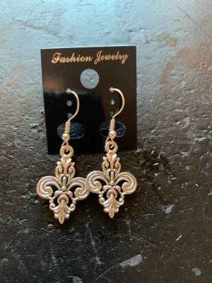 Silvriga örhängen med liljemotiv - Snygga silvriga örhängen med klassiskt liljemotiv och krokfäste. De har en detaljerad, ornamenterad design som ger en cool vintagekänsla. Perfekta för dig som gillar statement-smycken och vill sticka ut lite extra.