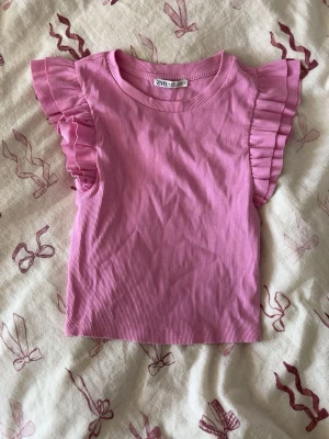 Rosa ribbad topp med volangärmar Zara - Supersöt rosa ribbad topp från Zara med volangdetaljer på ärmarna. Toppen är ärmlös med breda volanger som ger en trendig och lekfull look. Perfekt att styla med jeans eller kjol för en färgglad outfit.