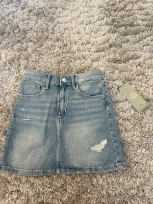 Jeanskjol  - Snygg blå jeanskjol i kort modell med slitna detaljer framtill. Kjolen har klassiska fickor fram och bak. Tillverkad i bomull och passar perfekt till en avslappnad stil. Aldrig använd och prislappen sitter kvar