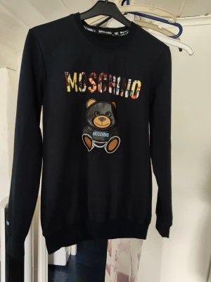 Svart Moschino tröja med björn - Cool svart tröja från Moschino med färgglad logga och en söt björn på bröstet. Tröjan har rund halsringning och långa ärmar. Perfekt för dig som gillar streetwear och vill sticka ut med en unik design. Kan inte garantera äkthet 