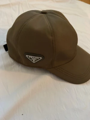 Olivgrön keps från Prada - Snygg olivgrön keps från Prada med klassisk böjd skärm och ikonisk metallogo framtill. Kepsen har sex paneler och justerbar rem baktill. Tillverkad i ett slätt, tåligt material som ger en exklusiv känsla.