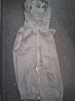 Grå ärmlös hoodie med dragkedja - Snygg grå ärmlös hoodie med dragkedja framtill och två fickor. Perfekt för lager-på-lager och chill stil. Mjuk bomullskänsla och huva med snörning för extra attityd. Fattas ett snöre som ska va i luvan 