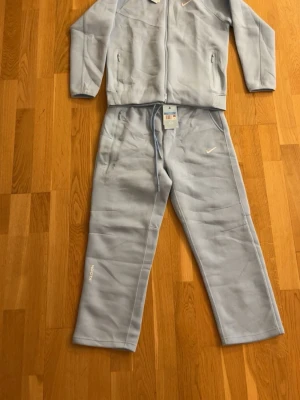 Ljusblått loungewear-set från Nike - Mysigt loungewear-set från Nike i ljusblått med dragkedjetröja och matchande byxor. Setet har diskreta Nike-loggor, dragkedjefickor på tröjan och dragsko i midjan på byxorna. Perfekt för chill dagar hemma. Materialet är mjukt och bekvämt. ‼️Pris kan diskuteras vid snabb affär‼️