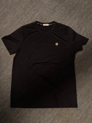 Svart Burberry t-shirt med logga - Snygg svart t-shirt från Burberry med rund hals och diskret Burberry-mönster i tyget. På bröstet sitter en liten rund logga i färg. Perfekt för dig som gillar stilrena plagg med exklusiv känsla. Storlek L men passar M 