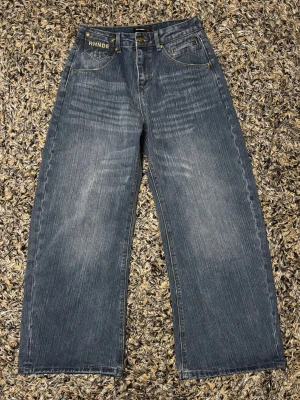 Vida jeans från RHNDE i blå denim - Säljer ett par blå jeans från RHNDE med bred passform och raka ben. Jeansen har klassiska fem fickor, guldfärgade detaljer och broderade bakfickor med R-logga. Tillverkade i denim med snygg tvättad look.