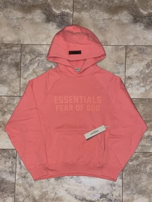 Koralrosa hoodie från Essentials fear of god  - Säljer en snygg korallrosa hoodie från Fear of God Essentials med tryckt logga på bröstet. Hoodien har huva, magficka och ribbade muddar. Perfekt för dig som gillar streetwear och vill sticka ut med en färgstark look. PASSAR INTE MIG FÖR STOR PÅ MIG