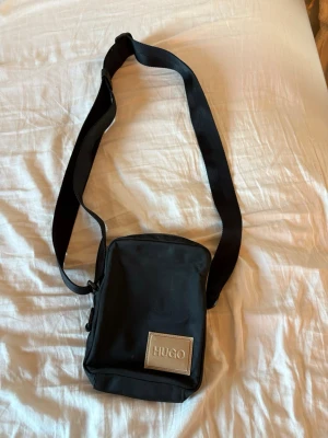 Svart axelväska från HUGO boss  - Snygg svart axelväska från HUGO med justerbar bred axelrem och en fyrkantig form. Väskan har en tydlig HUGO-logga framtill i silverfärg och är tillverkad i slitstarkt tygmaterial. Perfekt för att bära det viktigaste med stil.