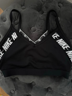 Svart sport-bh från Nike - Svart sport-bh från Nike med tunna axelband och vita logotyper längs kanterna. Bh:n har en sportig design med korsade band i ryggen och bred resår under bysten. Tillverkad i stretchigt material som ger bra stöd och komfort. Vet inte exakta storlek tyvärr men skulle säga passar som S. Den har inga inlägg! 