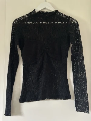 Svart spetsblus Nelly  - Snygg svart blus i genombruten spets med lång ärm och hög hals. Figurnära passform och transparent material ger en trendig look. Perfekt att styla lager på lager eller som statement-plagg. Storlek S FRÅN NELLY