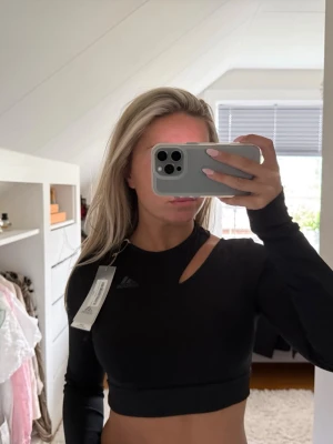 Svart croppad topp från Adidas - Snygg svart croppad topp från Adidas med lång ärm och unik cut out-detalj vid axeln. I stretchigt syntetmaterial som passar perfekt till träning eller en sportig look. Diskret Adidas-logga på bröstet och tight passform.