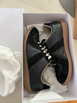 Maison Margiela gats - Svarta sneakers från Maison Margiela med ovandel i läder och mocka, vita snören och klassisk gummisula i brunt. Snygg retrostil med diskreta detaljer och låg profil. Perfekta för dig som gillar stilrena och tidlösa sneakers.skorna är såklart äkta fås med låda dustbag kort.