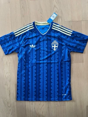 Blå Sverige fotbollströja Adidas - Säljer en blå Sverige fotbollströja från Adidas med gult och blått mönster, gula ränder på axlarna och broderad landslagssköld. Tröjan har korta ärmar, V-ringning och är tillverkad i ett lätt, ventilerande material. Perfekt för match eller träning.