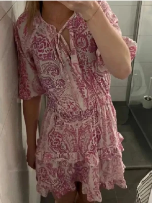 Rosa och vit kortklänning från Zara - Supersöt kortklänning från Zara i rosa och vitt med blommigt mönster. Klänningen har volangdetaljer, knytning vid halsen och trekvartsärmar. Perfekt för varma dagar och har en luftig passform.