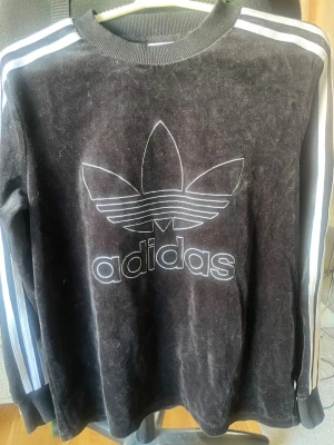 Svart Adidas tröja med vita ränder - Svart långärmad Adidas tröja med klassiska vita ränder längs ärmarna och stort vitt Trefoil-logo tryck på bröstet. Rund halsringning och ribbade muddar. Mjuk och lite glansig materialkänsla, perfekt för en sportig och avslappnad stil. Bomull och polyester