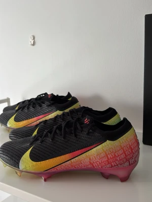 Nike Mercurial VINI VOA VINI FLY (Limited Edition) - Snygga Nike Mercurial fotbollsskor i svart med stora gula och röda detaljer längs sidorna. Skorna har en låg profil, snörning och textilöverdel med struktur för bättre bollkontroll. Sulan är rosa och designad för bra grepp på planen. Perfekta för dig som vill sticka ut på matchen.