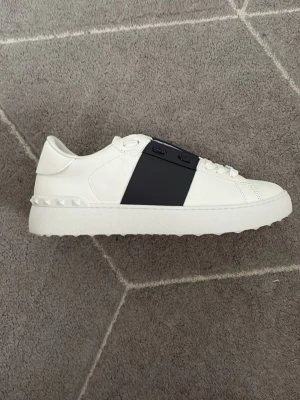 Valentino Garavani vita sneakers - Snygga vita sneakers från Valentino Garavani med en bred navy rem över mitten. Skorna är i läder och har coola nitar på hälen. Perfekta för dig som gillar stilrena och exklusiva detaljer.