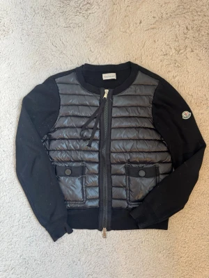 Svart cardiganjacka från Moncler - Snygg svart cardiganjacka från Moncler med quiltad framsida i glansigt material och stickade ärmar. Jackan har två fickor framtill med knappar och dragkedja hela vägen. Klassisk Moncler-logga på ärmen och rund halsringning.