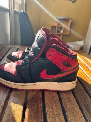 Air Jordan 1 Mid Svart/Röd - Säljer ett par Air Jordan 1 Mid sneakers i svart och rött med klassisk Nike Swoosh och Jordan Wings-logga på sidan. Skorna har en vit mellansula och röd yttersula. Ovandelen är i läder och textil, och modellen har en ikonisk siluett som alltid är trendig.