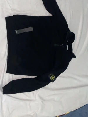 Svart hoodie från Stone Island - Svart hoodie från Stone Island med klassisk logotyp-patch på vänster ärm. Hoodien har huva och ribbade muddar. Perfekt för en streetwear-look och tillverkad i mjukt material för extra komfort.