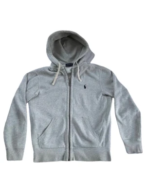 Polo Ralph Lauren Zip Sweater - Snygg grå hoodie från Polo Ralph Lauren med dragkedja, huva med dragsko och två fickor framtill. Klassisk logga broderad på bröstet. Perfekt för en avslappnad och stilren look.