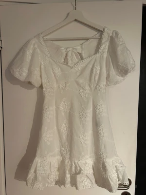Vit blommig kortklänning med volang - Superfin vit kortklänning med broderade blommor, puffärmar och volangkant nertill. Klänningen har v-ringning och en stor rosett i ryggen som detalj. Perfekt för sommarens alla tillfällen!