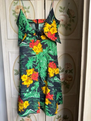 Blommig sommarklänning med smala axelband - Färgstark klänning med tropiskt blommönster i gult, rött och grönt på svart botten. Klänningen har smala axelband, omlott-detalj framtill och lätt utställd kjol. 