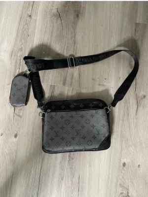  grå axelväska med monogram - Snygg grå axelväska med klassiskt monogrammönster i svart. Väskan har justerbar svart axelrem och en mindre avtagbar ficka. Tillverkad i canvas med detaljer i läder och silverfärgade metalldelar.