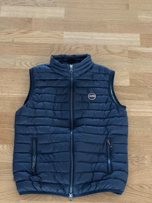 Mörkblå dunväst från Colmar - Snygg mörkblå dunväst från Colmar med quiltad design och två dragkedjefickor framtill. Västen har hög krage och en diskret logotyp på bröstet. Insidan är ljusgrön för en cool kontrast. Perfekt för lager-på-lager-stil. 