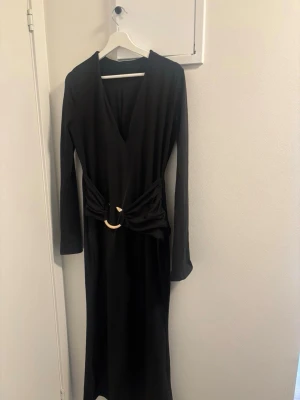 Svart långklänning med v-ringning - Stilren svart långklänning med djup v-ringning och långa ärmar. Klänningen har en snygg detalj i midjan med ett dekorativt bälte och guldfärgat spänne. Perfekt för dig som gillar minimalistisk och elegant stil. Ordinarie pris 400